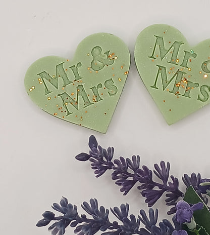 Two green heart-shaped decorations with 'Mr & Mrs' text, accompanied by purple floral elements on a light gray background.