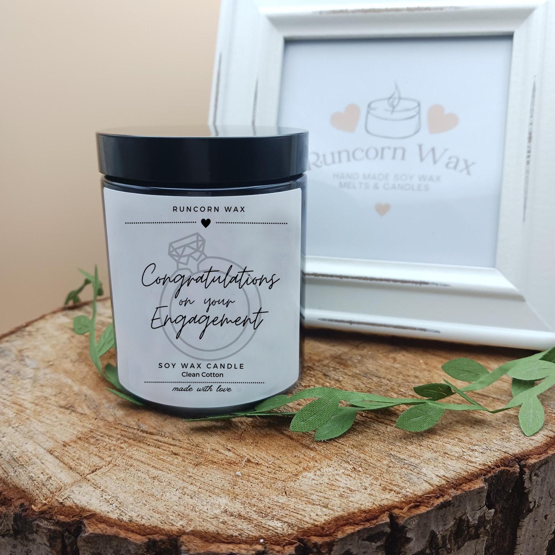 Soy wax candle with 'Congratulations on your Engagement' text on a wooden surface, framed picture in the background.