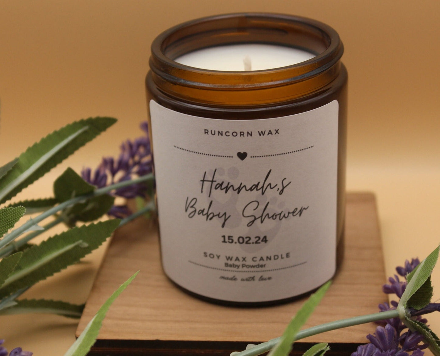 Candle in an amber jar with a personalized label on a wooden surface, surrounded by lavender flowers.