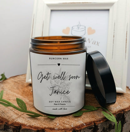 Scented candle with personalized label on a wooden surface, framed picture in the background get well soon