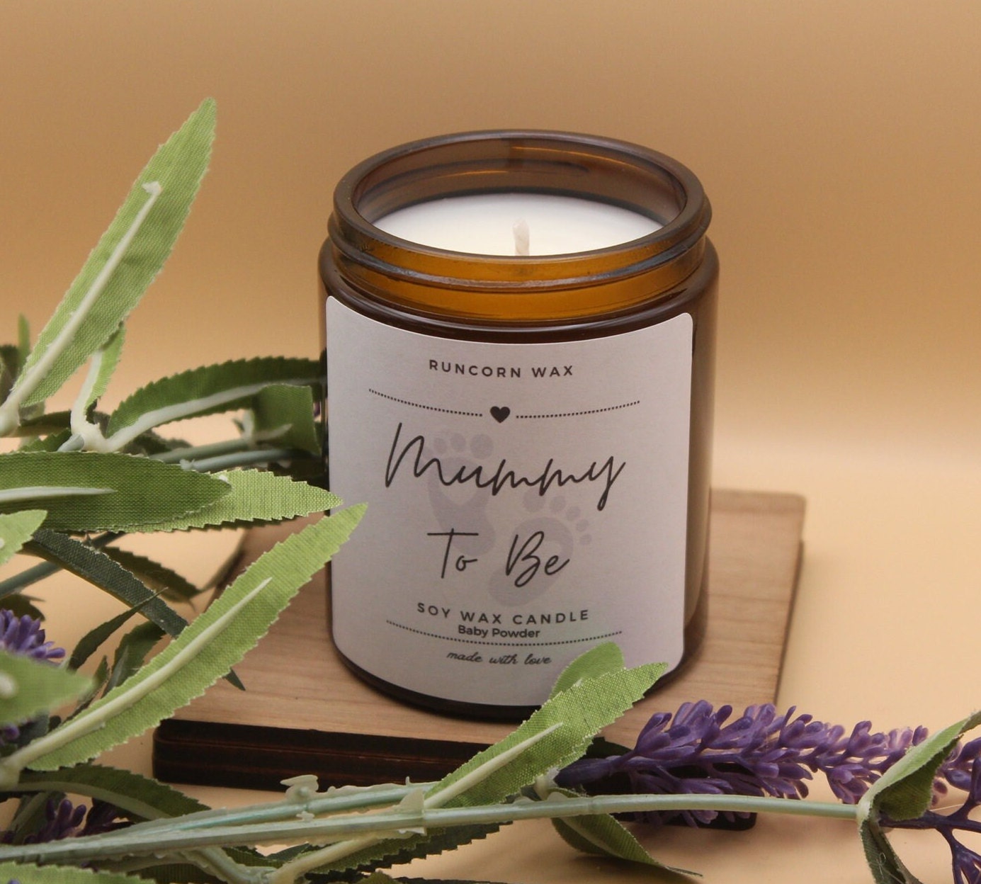 Candle labeled 'Mummy To Be' with lavender and green leaves on a wooden surface