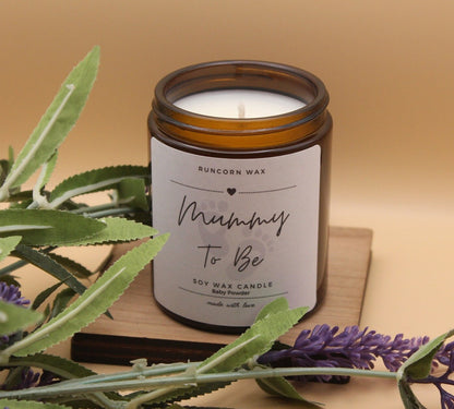 Candle labeled 'Mummy To Be' with lavender and green leaves on a wooden surface