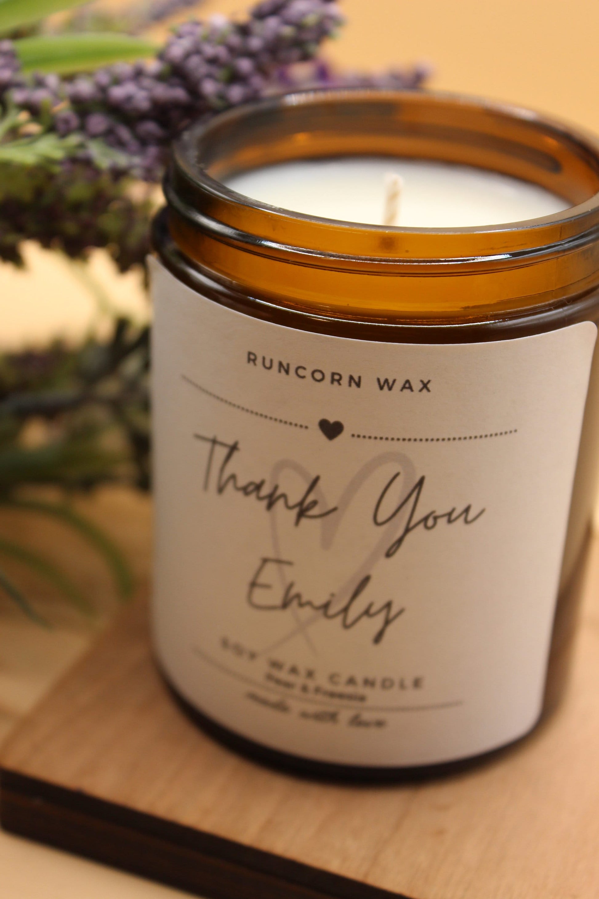 Candle in a glass jar with a 'Runcorn Wax' label on a wooden surface with lavender flowers.