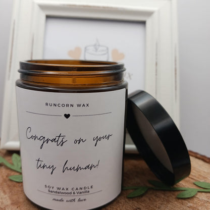Candle with a label reading 'Congrats on your tiny human!' by Runcorn Wax, sitting on a wooden surface with green leaves.