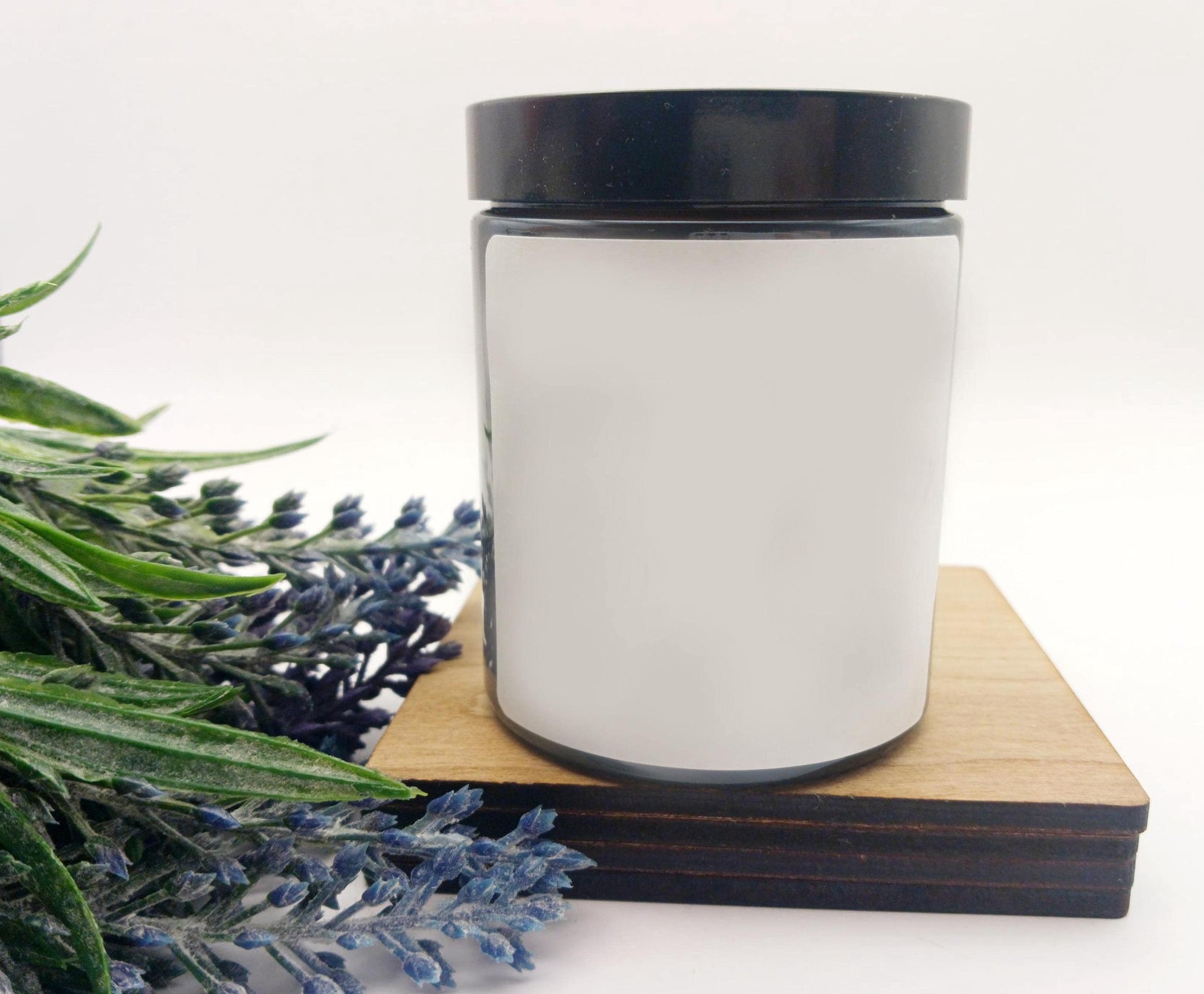 White candle jar with black lid on a wooden coaster, surrounded by lavender flowers.
