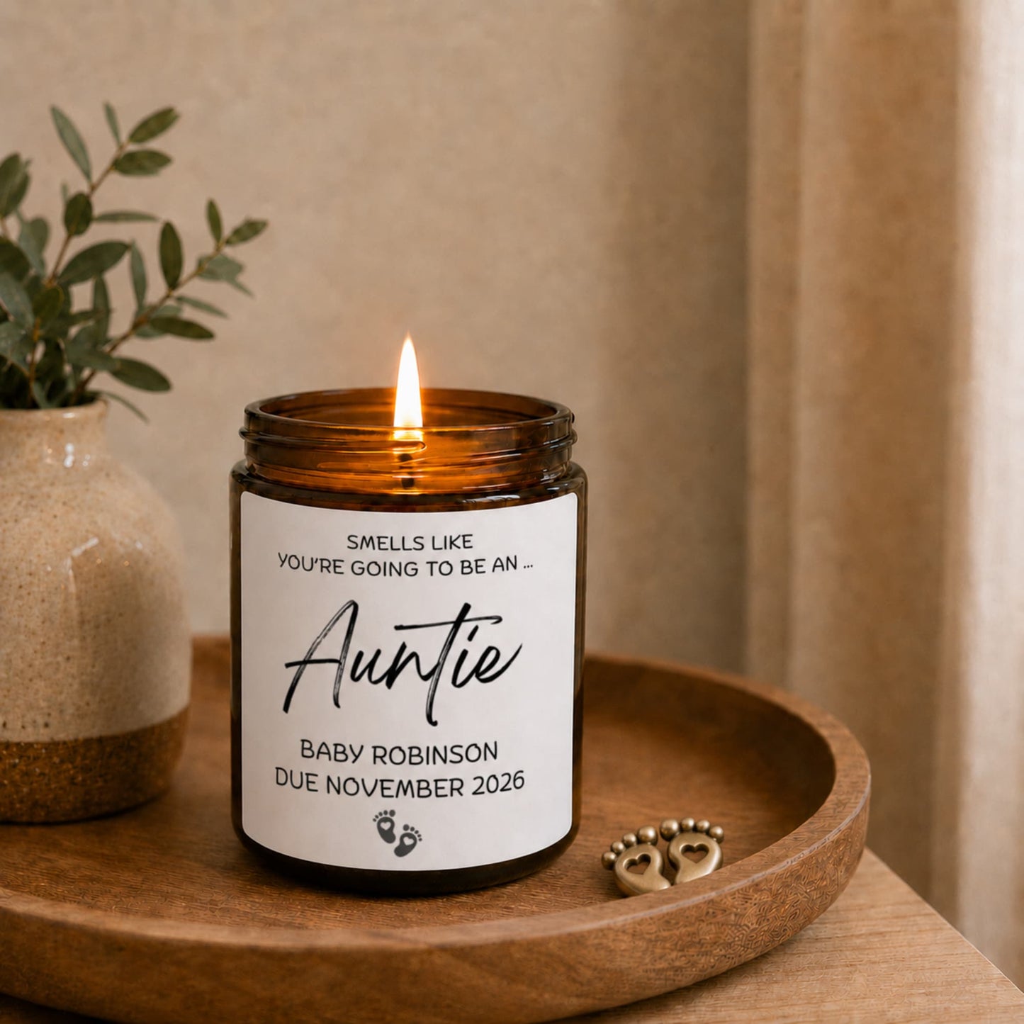 Personalised auntie pregnancy announcement candle in amber glass jar, lit on wooden tray with baby feet charm, Baby Robinson due November 2026 gift. Candle in a jar with a personalized label on a wooden tray, next to a plant.
