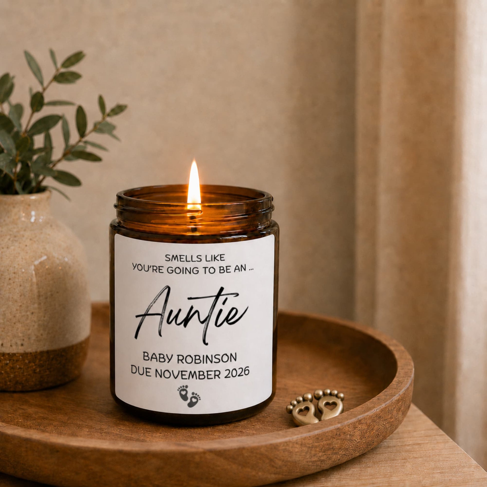 Personalised auntie pregnancy announcement candle in amber glass jar, lit on wooden tray with baby feet charm, Baby Robinson due November 2026 gift. Candle in a jar with a personalized label on a wooden tray, next to a plant.