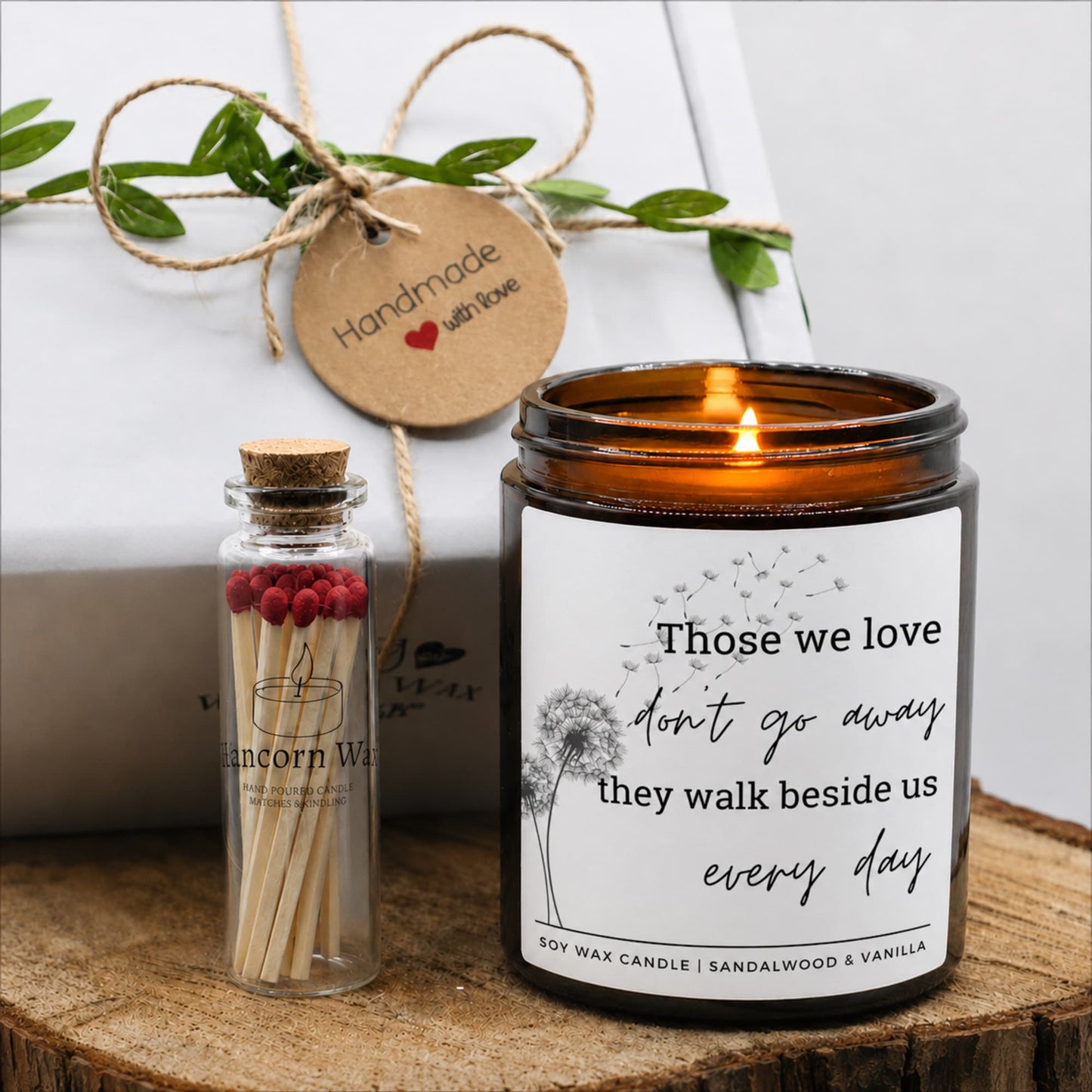 An amber glass jar candle with a white printed label reading 'Those we love don't go away they walk beside us every day' is lit on a wooden surface. A small glass match bottle with red matches and a cork stopper sits beside it. bereavement loss sympathy candle gift