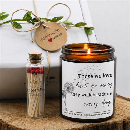 An amber glass jar candle with a white printed label reading 'Those we love don't go away they walk beside us every day' is lit on a wooden surface. A small glass match bottle with red matches and a cork stopper sits beside it. bereavement loss sympathy candle gift