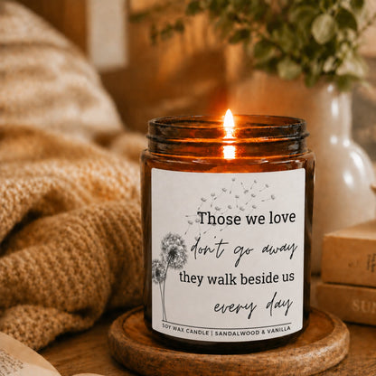 A lit amber glass jar candle sits on a wooden coaster on a rustic table, surrounded by cozy knitted blankets, books, and a vase with greenery. The white label features a dandelion illustration and the text: 'Those we love don't go away they walk beside us every day' and 'SOY WAX CANDLE | SANDALWOOD & VANILLA'. bereavement loss sympathy candle gift