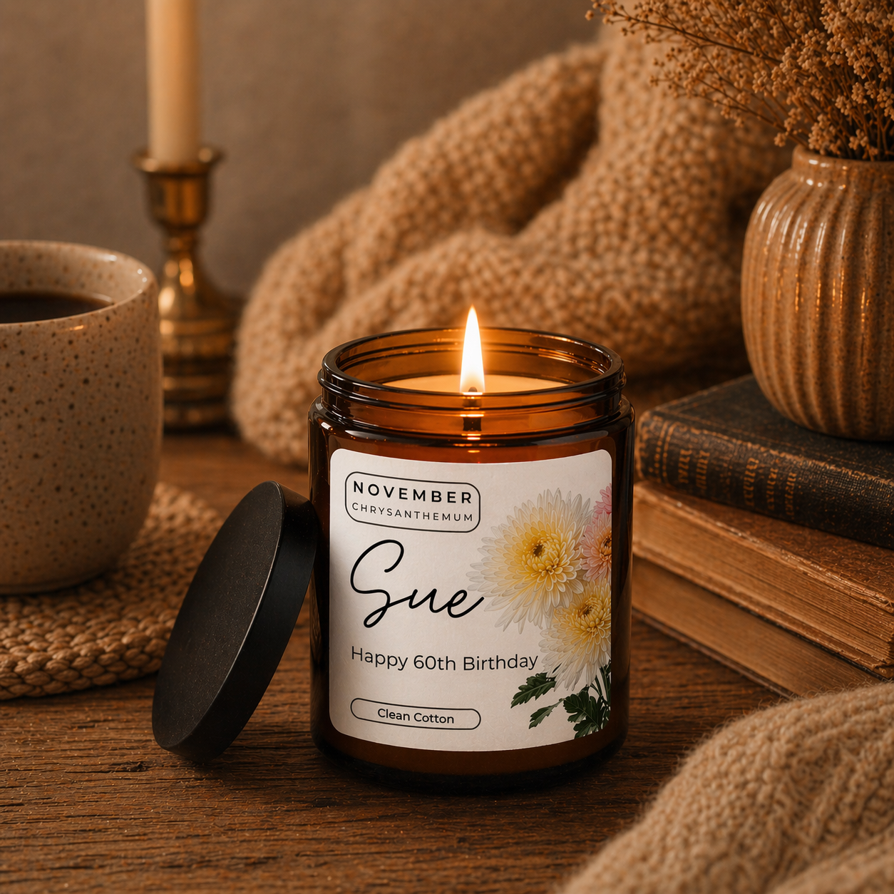 A warm, cozy scene featuring a lit amber glass candle on a rustic wooden table. The candle label reads “November Chrysanthemum – Sue – Happy 60th Birthday – Clean Cotton,” decorated with soft floral illustrations. Surrounding the candle are a ceramic mug, stacked vintage books, a knitted blanket, and a vase with dried flowers, all softly lit by a golden, ambient glow.