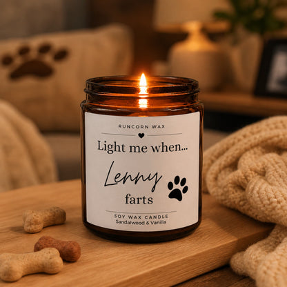 Lit amber glass candle with “Light me when Lenny farts” label, styled in a warm cosy home setting with a knitted blanket and dog treats nearby.