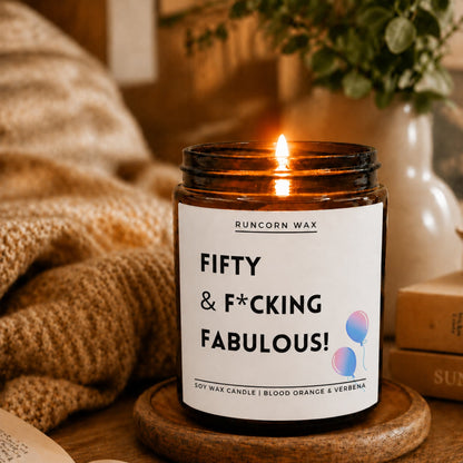 A lit soy wax candle in an amber glass jar sits on a wooden coaster on a table, surrounded by a cozy knitted blanket, books, and a vase with greenery. The label reads 'RUNCORN WAX' and 'FIFTY & *F*CKING FABULOUS!'.