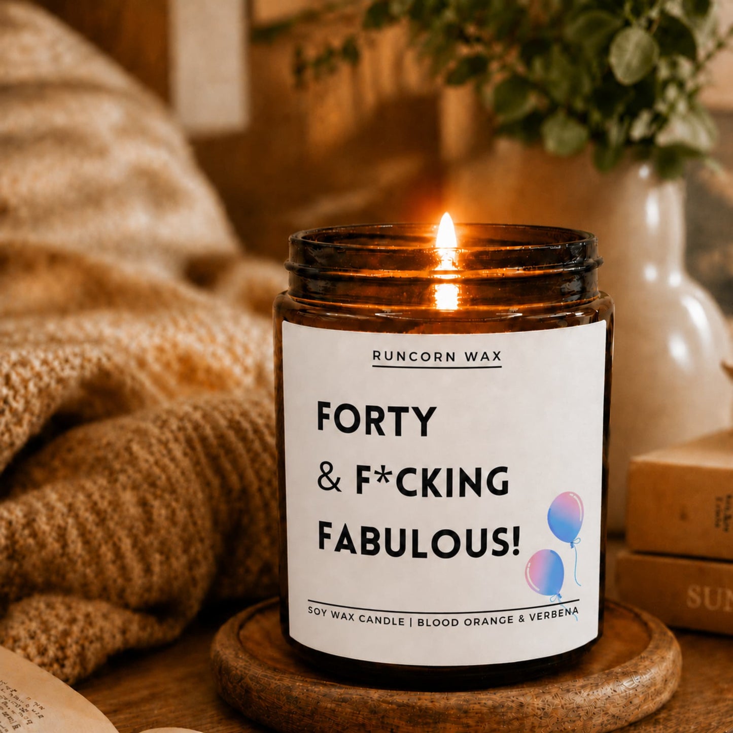 A lit soy wax candle in an amber glass jar sits on a wooden coaster in a cozy indoor setting. The white label reads 'RUNCORN WAX' and 'FORTY & F*CKING FABULOUS!' with small balloon graphics.