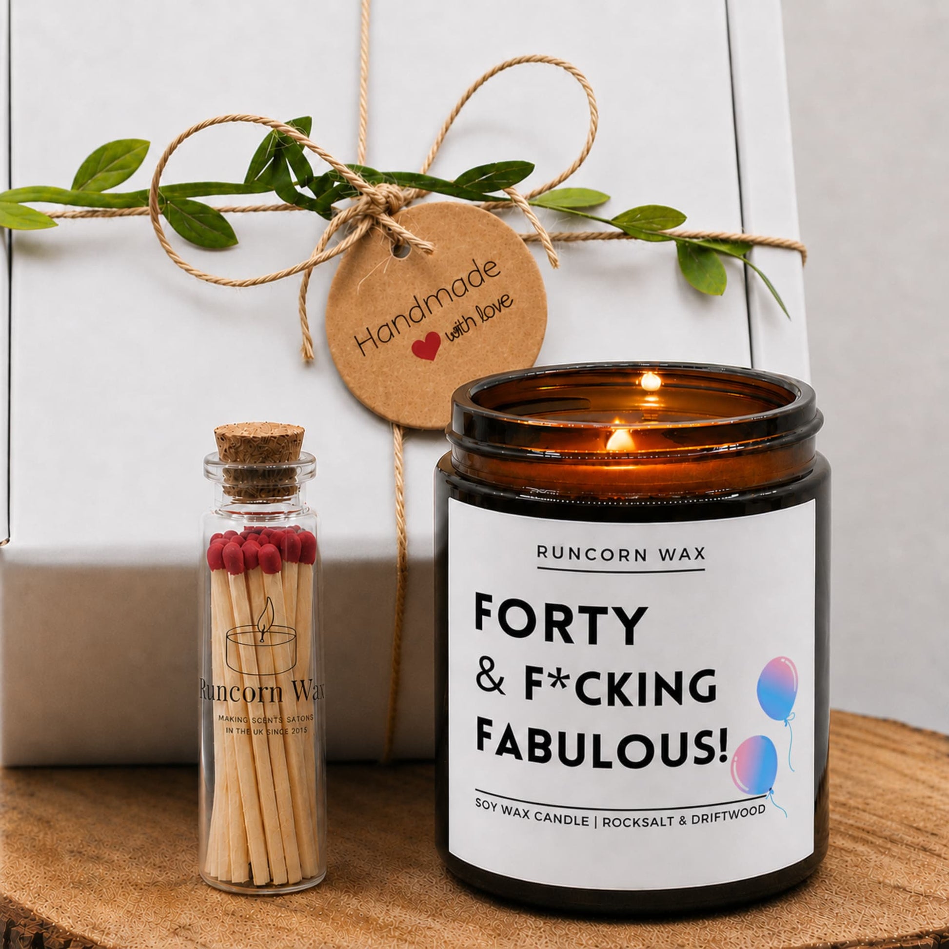 An amber glass jar candle labeled 'FORTY & F*CKING FABULOUS!' by Runcorn Wax sits on a wooden surface next to a small glass match bottle with lit matches. A white gift box wrapped with twine and a round kraft tag reading 'Handmade with love' is in the background.