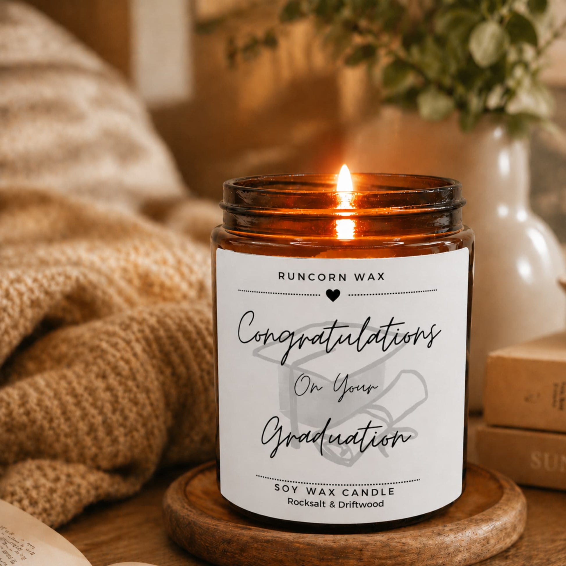 Graduation-themed candle with a label on a wooden surface, surrounded by cozy decor.