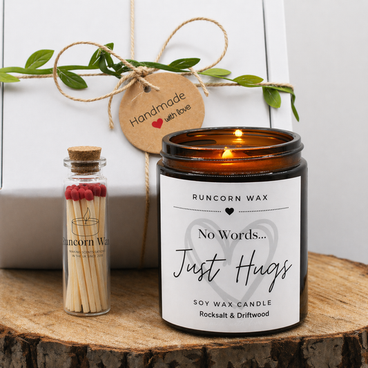 Candle labeled 'No Words... Just Hugs' by Runcorn Wax on a wooden surface with a bottle of matches and greenery.