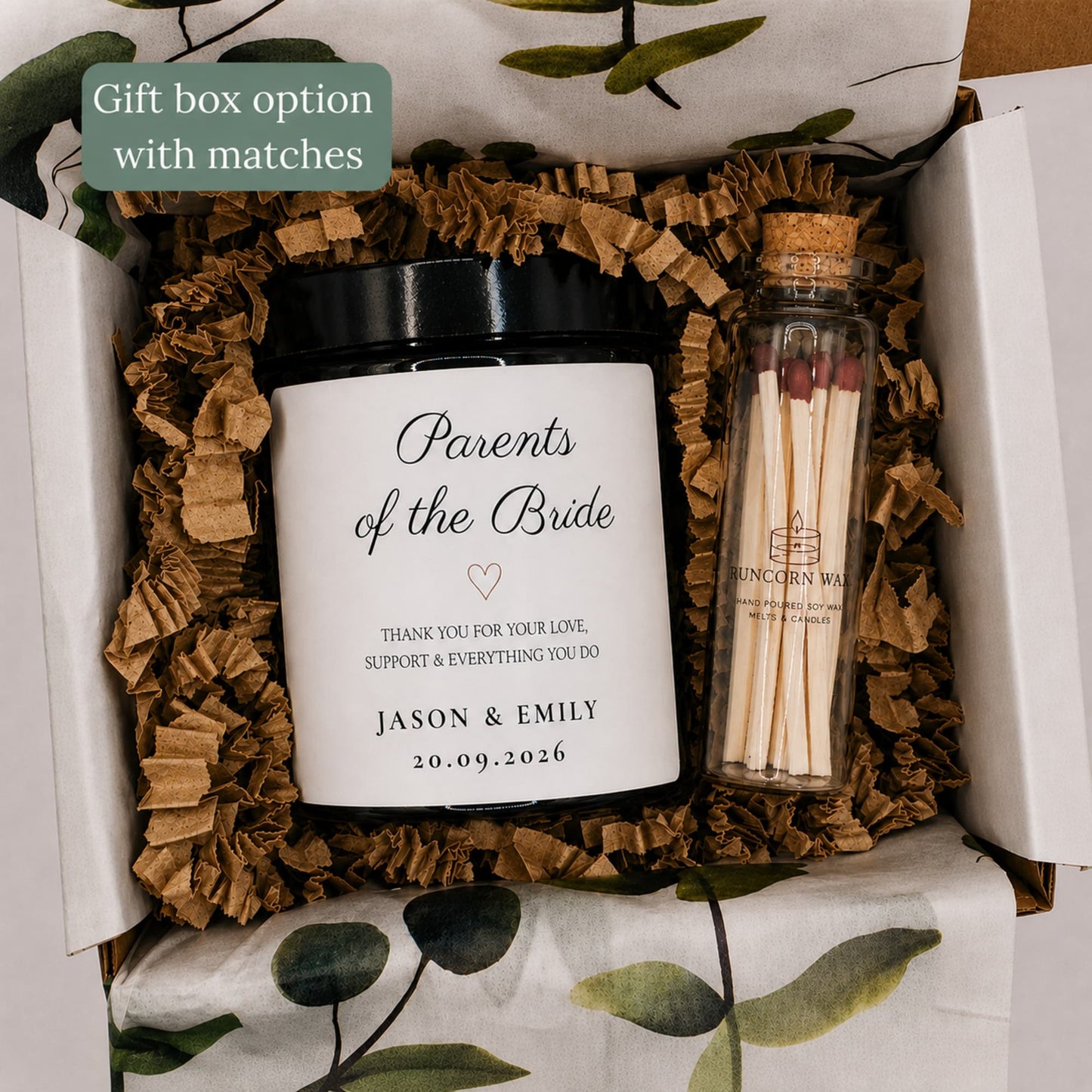 A personalized gift box set containing a white jar candle with a black lid labeled 'Parents of the Bride' and a small glass match bottle with wooden matches. The items are arranged in brown crinkle paper filler inside an open white box with green leaf-patterned tissue paper.