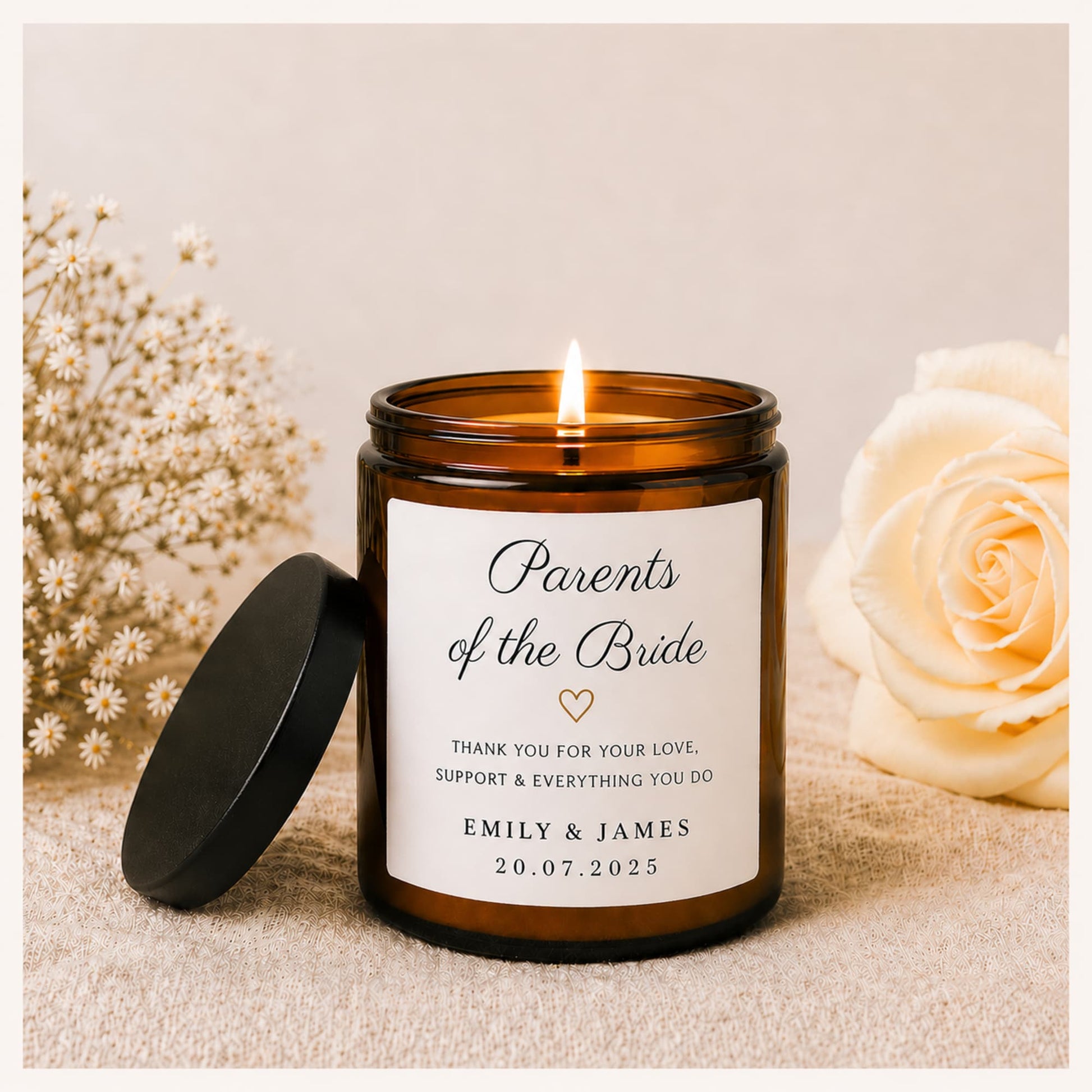 A personalized amber glass candle jar with a black lid resting beside it, lit and set on a neutral fabric surface with dried baby's breath and a pale rose in the background. The front label reads 'Parents of the Bride' with the names 'EMILY & JAMES' and a date.