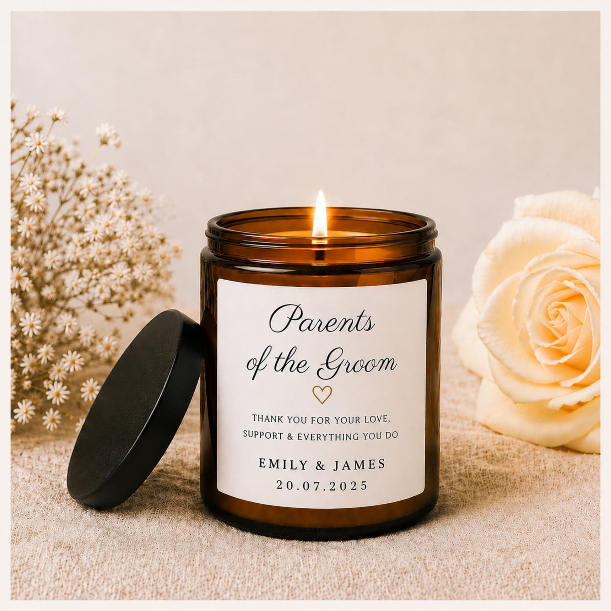 A lit amber glass jar candle with a black lid resting beside it on a beige fabric surface. The front label reads 'Parents of the Groom' with a small heart and personalized text for Emily & James, 20.
