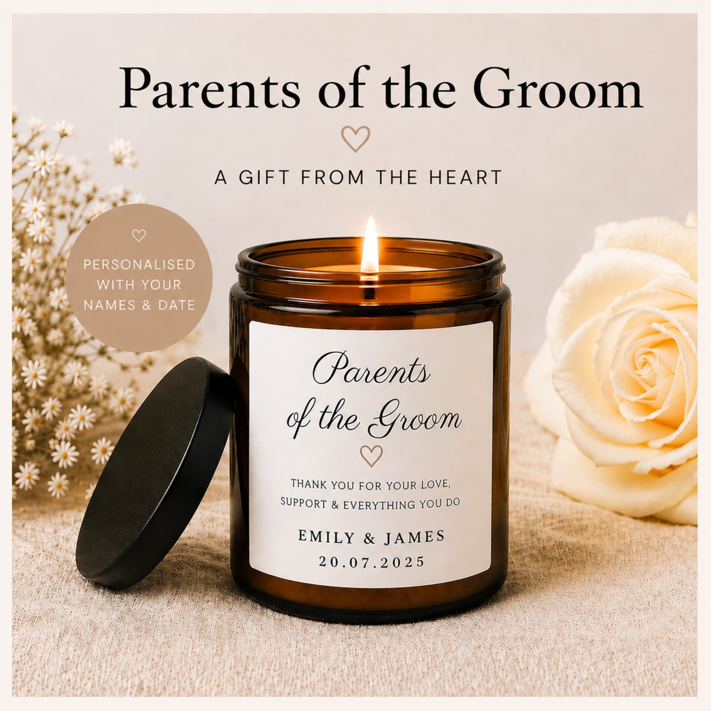 A personalized amber glass jar candle with a black lid resting beside it, lit and centered on a neutral fabric surface. Large cream roses and small white flowers decorate the background, with text reading 'Parents of the Groom' and 'A Gift from the Heart.