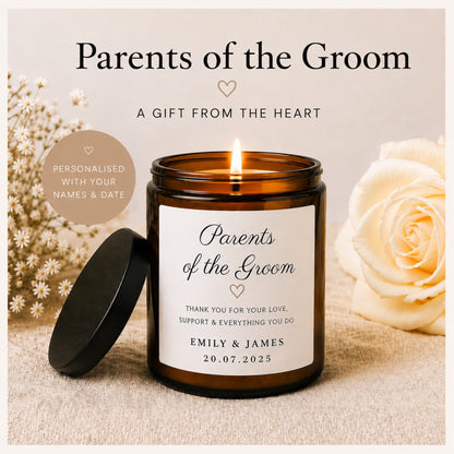 A personalized amber glass jar candle with a black lid resting beside it, lit and centered on a neutral fabric surface. Large cream roses and small white flowers decorate the background, with text reading 'Parents of the Groom' and 'A Gift from the Heart.