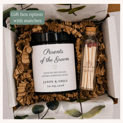 A personalized gift box set containing a black jar candle with a white label reading 'Parents of the Groom' and a small glass matchbox with wooden matches. The items are arranged in brown crinkle paper filler inside a white box with green leaf-patterned tissue paper.