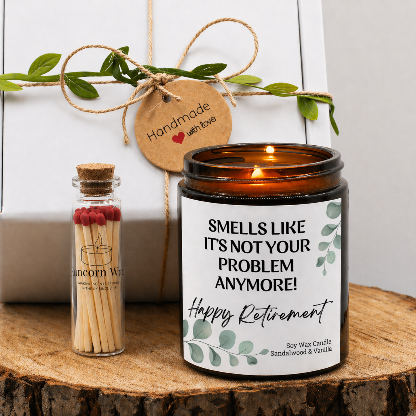 Soy wax retirement candle with “Smells Like It’s Not Your Problem Anymore – Happy Retirement” label, amber jar on wooden surface with match bottle and gift box.