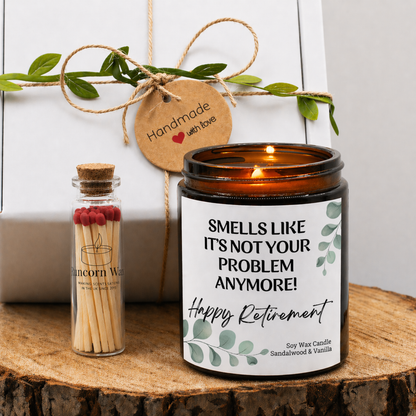 Soy wax retirement candle with “Smells Like It’s Not Your Problem Anymore – Happy Retirement” label, amber jar on wooden surface with match bottle and gift box.