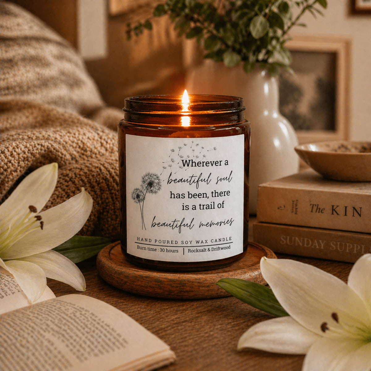 A lit amber glass jar candle sits on a wooden coaster in a cozy styled setting with books, lilies, and a knitted blanket. The label reads, 'Wherever a beautiful soul has been, there is a trail of beautiful memories' and identifies the brand as Rocknroll & Dirtwood.