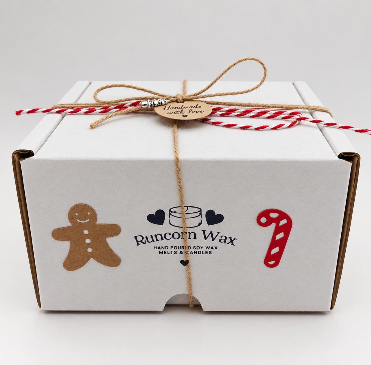 A white gift box tied with brown twine and red and white twine. The box has a gingerbread man sticker, a candy cane sticker, and a sticker that says "Runcorn Wax Hand Made Soy Wax Melts & Candles".