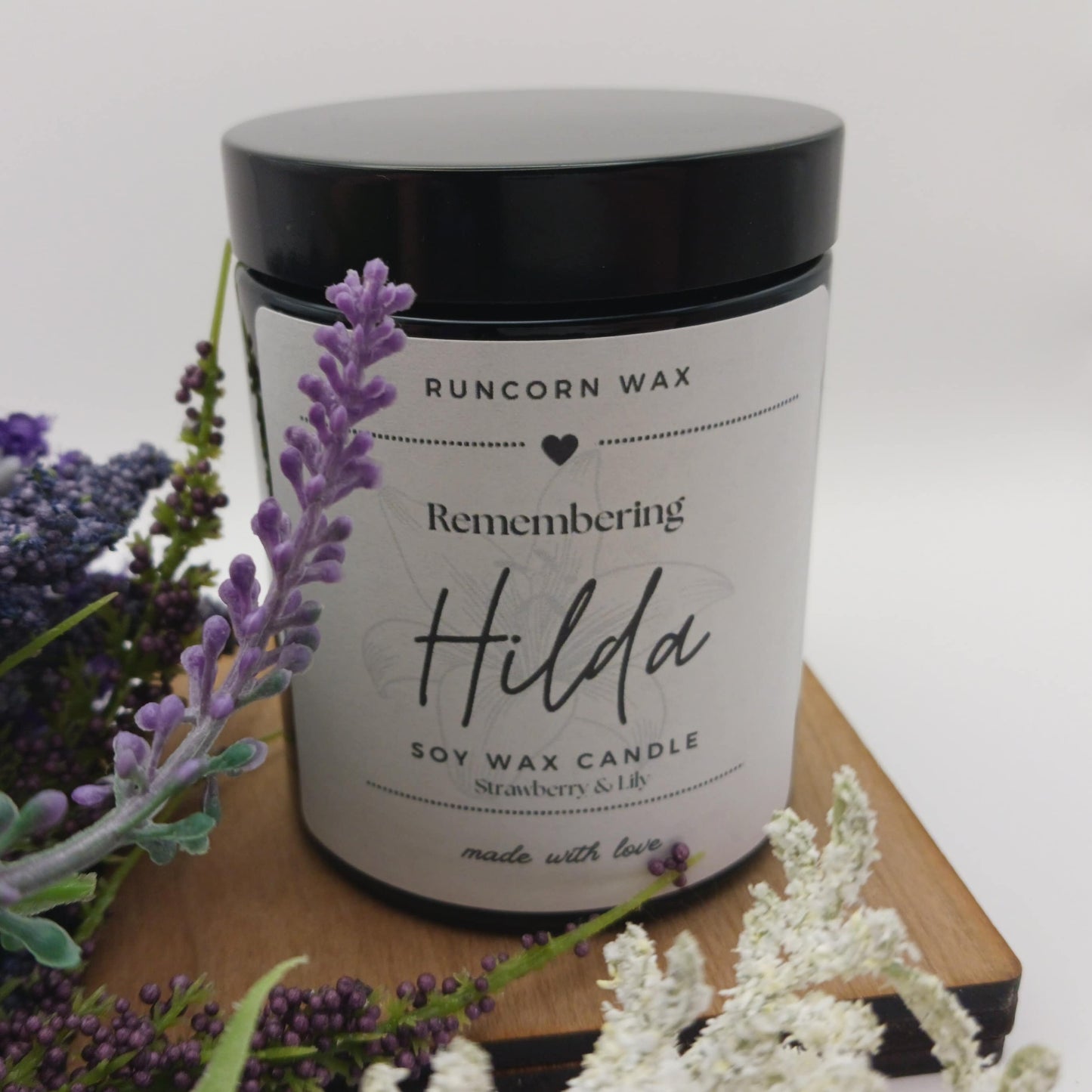 Runcorn Wax candle labeled 'Remembering Hilda' with lavender and white flowers on a wooden surface.
