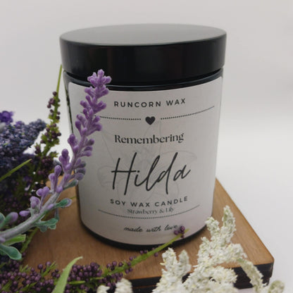 Runcorn Wax candle labeled 'Remembering Hilda' with lavender and white flowers on a wooden surface.