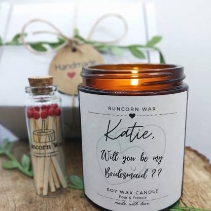 Candle with 'Katie, Will you be my Bridesmaid?' message and a small bottle of matches on a wooden surface.