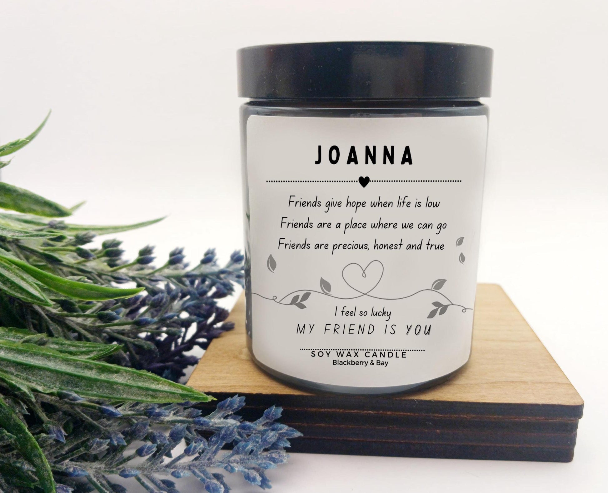 Candle with a label reading 'JOANNA' and text, surrounded by lavender on a white background