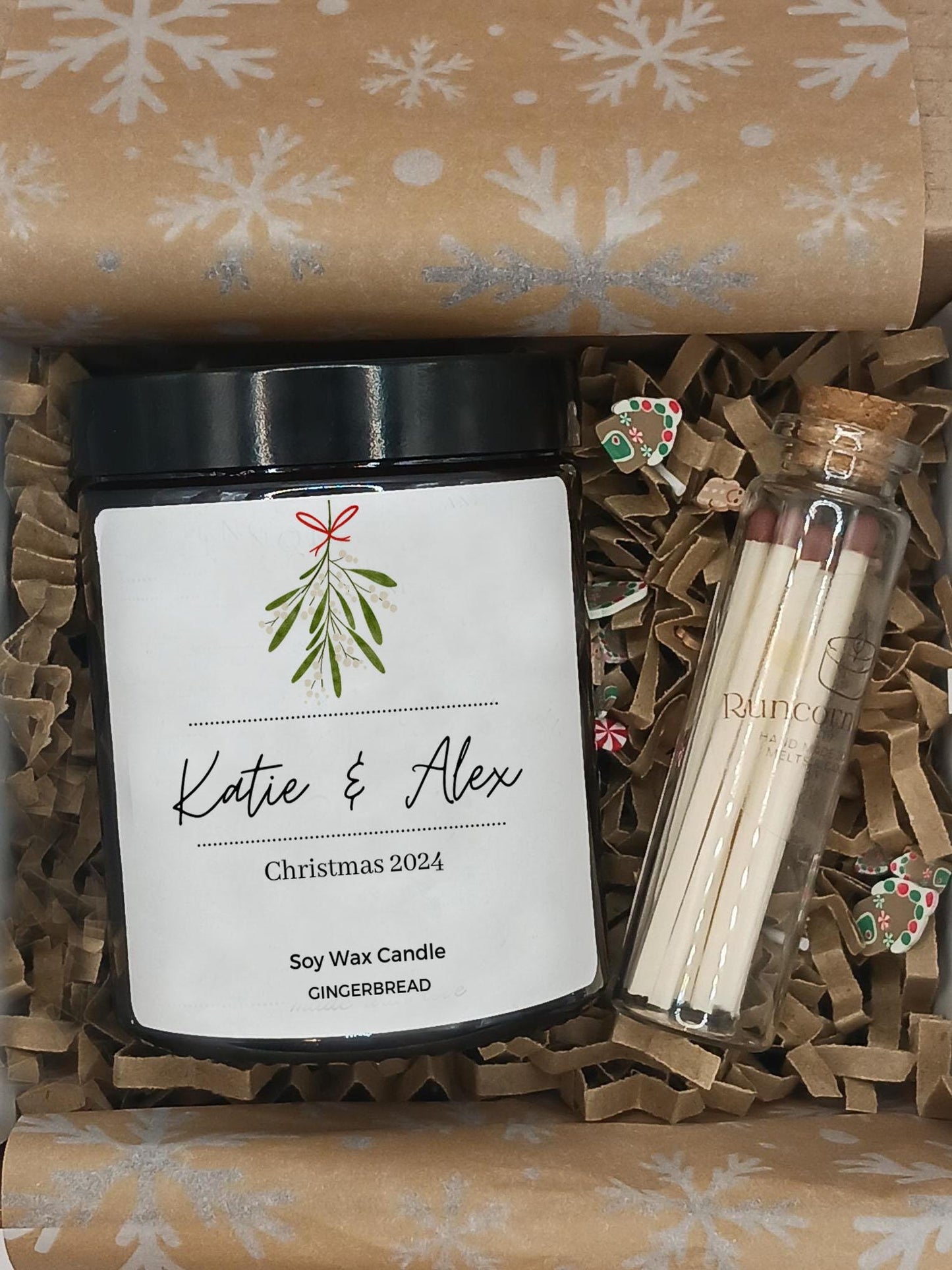 Gift set with a candle labeled 'Katie & Alex Christmas 2024', matches, and decorative items in a box with snowflake pattern.