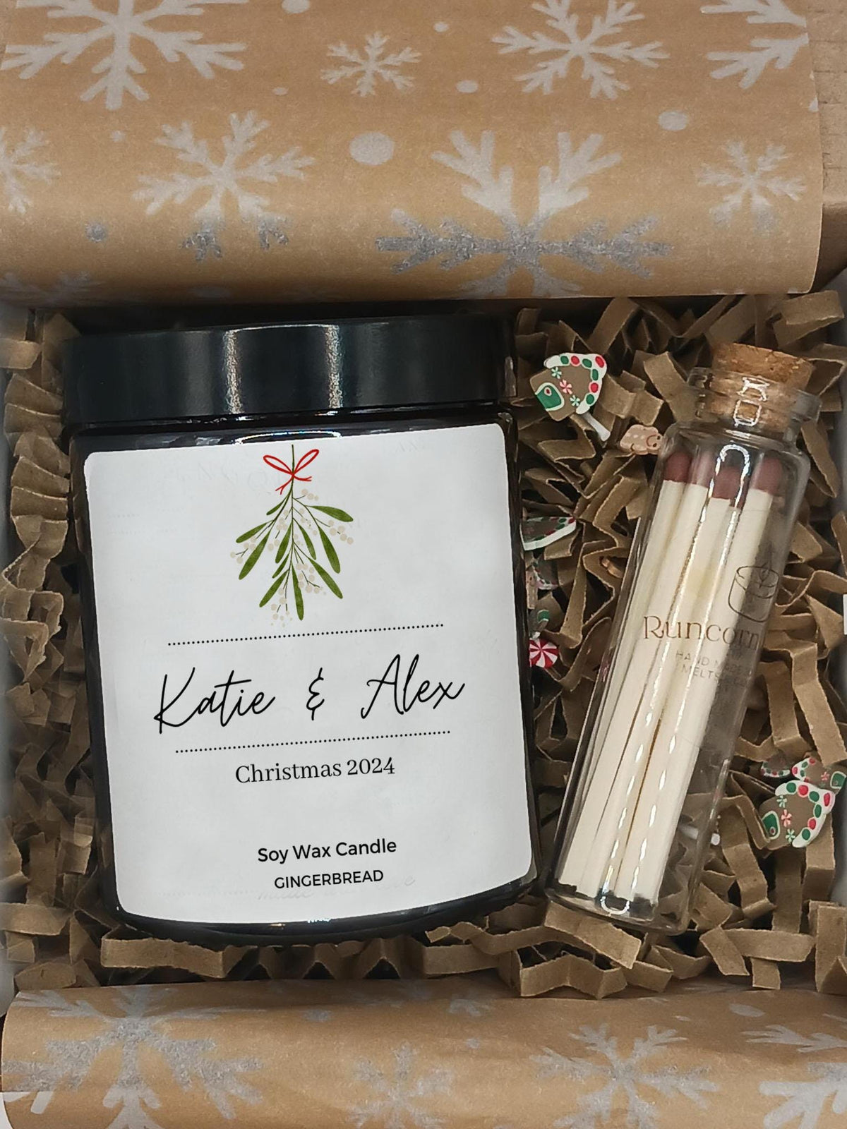 Gift set with a candle labeled 'Katie & Alex Christmas 2024', matches, and decorative items in a box with snowflake pattern.
