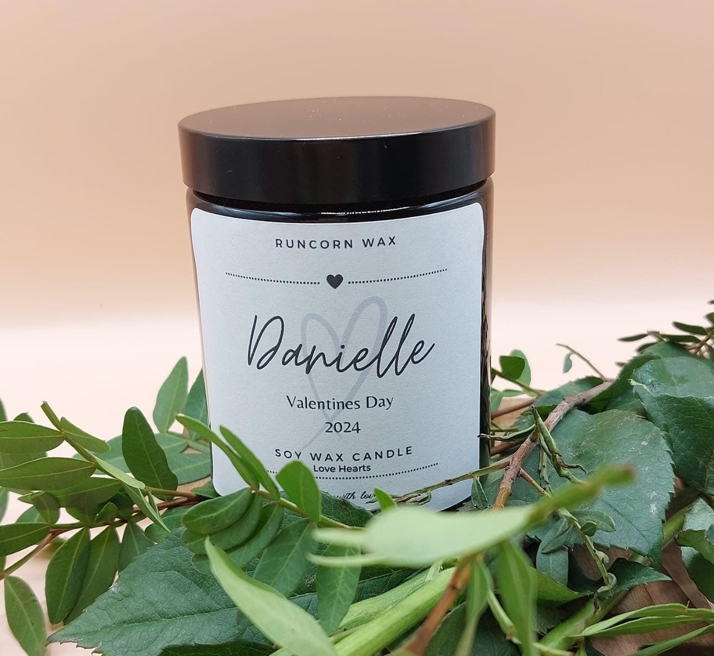 Candle labeled 'Danielle' with a heart design, surrounded by green leaves on a beige background