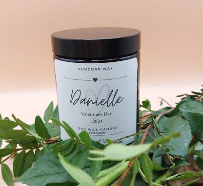 Candle labeled 'Danielle' with a heart design, surrounded by green leaves on a beige background