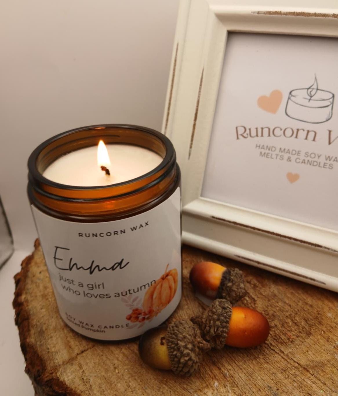 Candle in an amber glass jar labeled 'Emelie' with a wooden frame and acorns in the background.