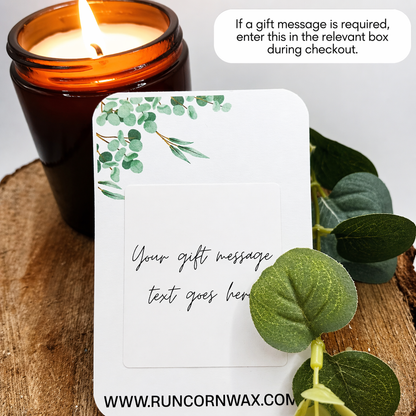 Close-up of a lit amber glass candle on a wooden surface beside a botanical-themed gift message card. The card features soft green leaf illustrations and placeholder text for a personalised message, with a leafy stem in the foreground. In the top corner, a note reads that gift messages can be entered during checkout.