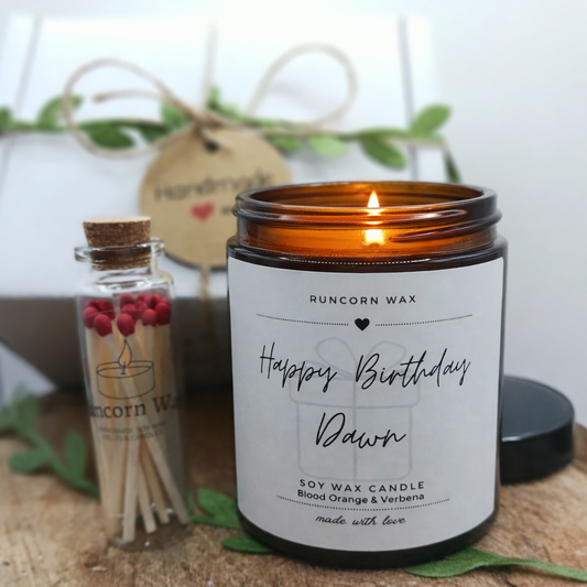 Candle in an amber jar with a label, matches, and decorative elements on a wooden surface, reading happy birthday personalised