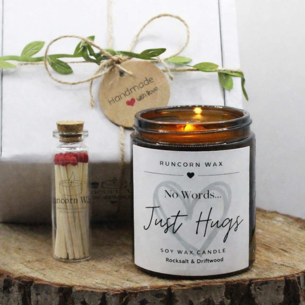 Candle labeled 'No Words... Just Hugs' by Runcorn Wax on a wooden surface with a bottle of matches and greenery.